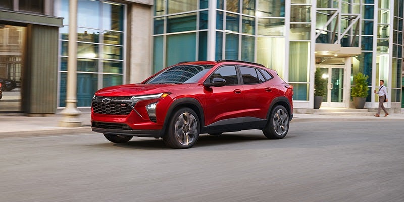 A red Chevrolet Trax drives on a city street past modern buildings, with a person walking on the sidewalk.
