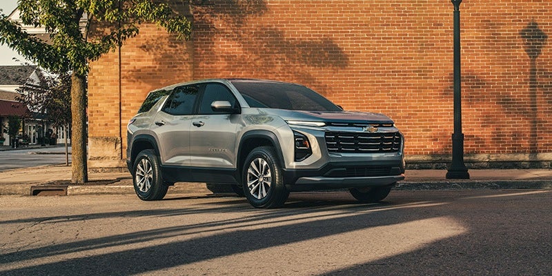 Silver Chevrolet Trax SUV parked on a street with a brick wall behind it and a tree to the left.